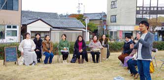 屋外で、椅子に座った人たちの横に立って話す様子